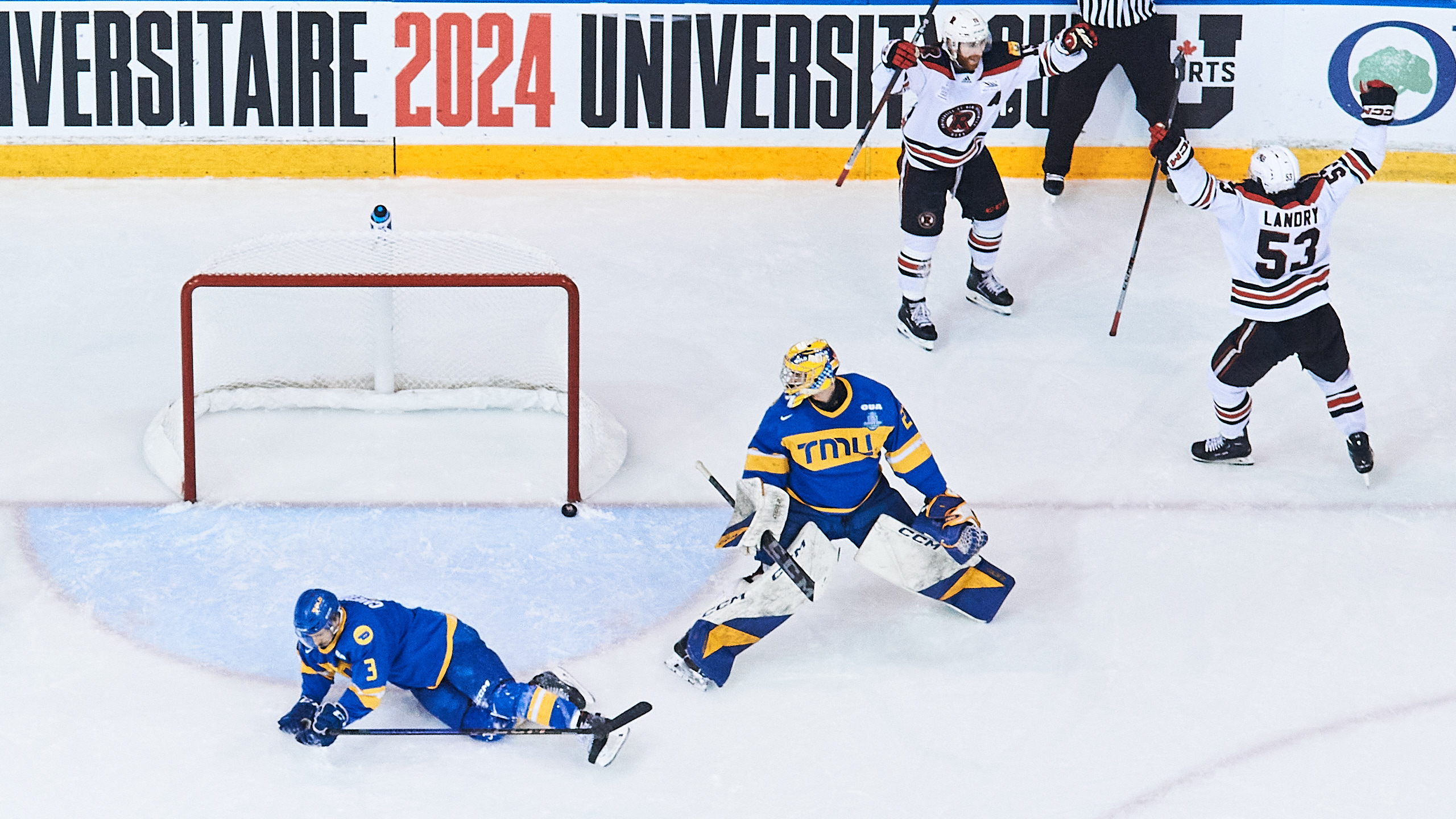A TMU player and the TMU goalie look on as a puck goes into the TMU net while UNB players celebrate