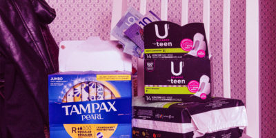 A stack of pads and tampons sit on a table with cash peeking out of one of the pad boxes.